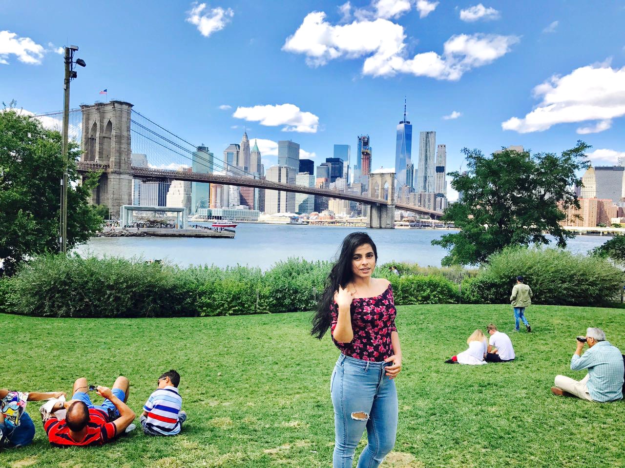 Pratima standing on a lawn at Brooklyn Bridge Park with the Brooklyn Bridge and Manhattan skyline behind her.