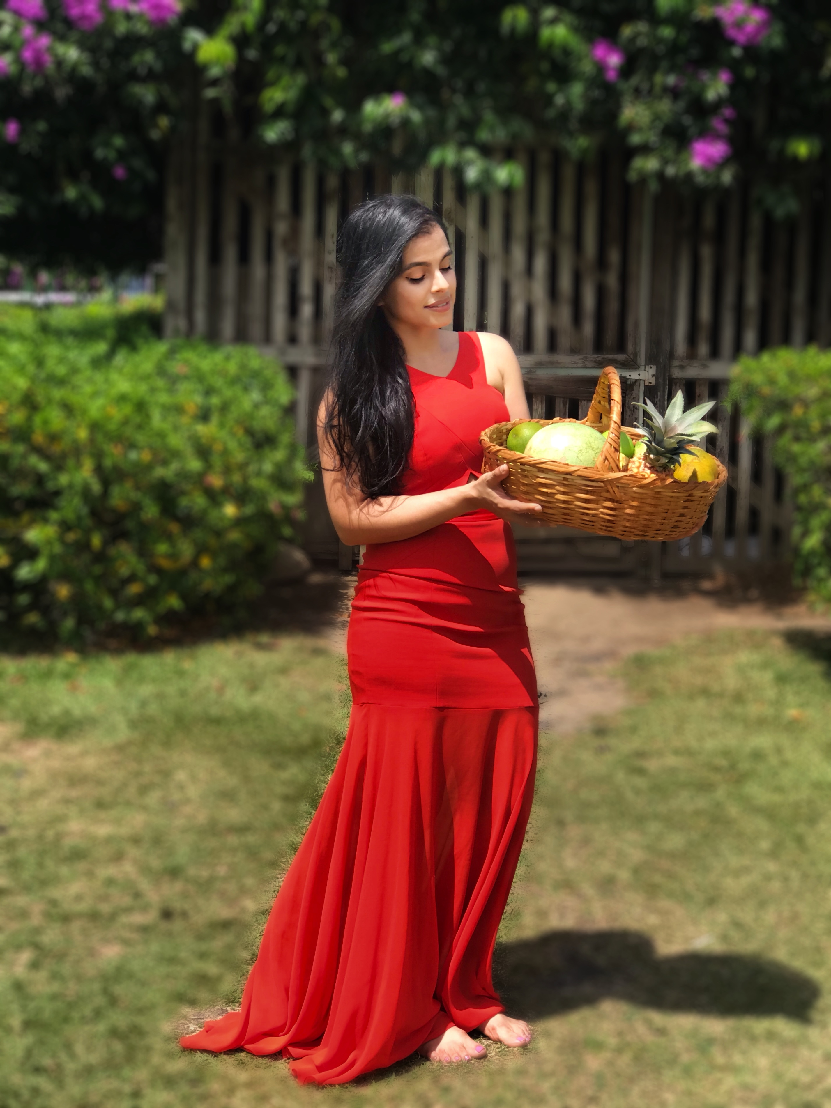Pratima holding a basket of fresh fruit outdoors in a red dress.