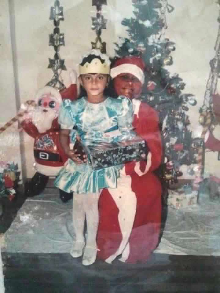 Child dressed in a festive costume sitting with Santa beside a decorated Christmas tree at a nursery-school Christmas party.